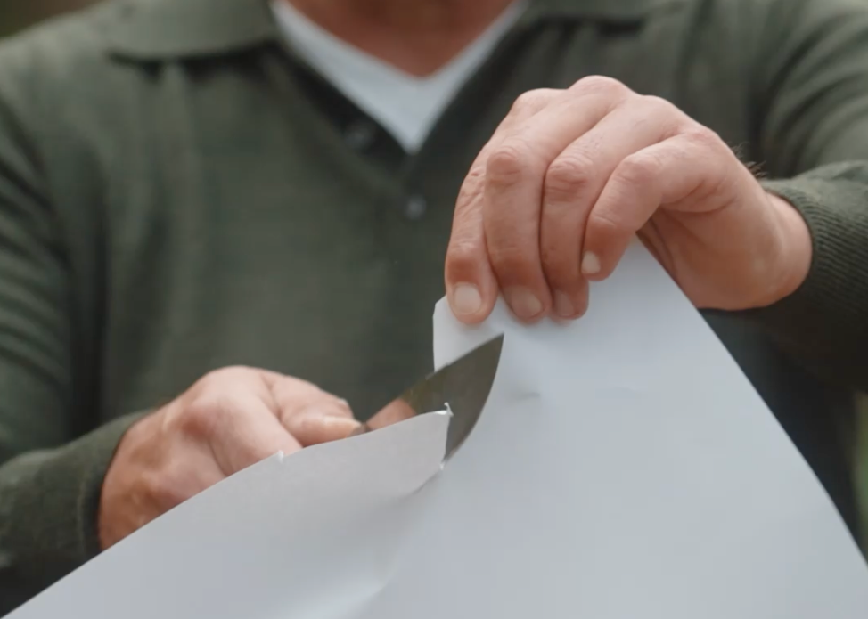 Eine Person, die ein weißes Blatt Papier mit einem Messer zerschneidet und einen grünen Pullover trägt.
